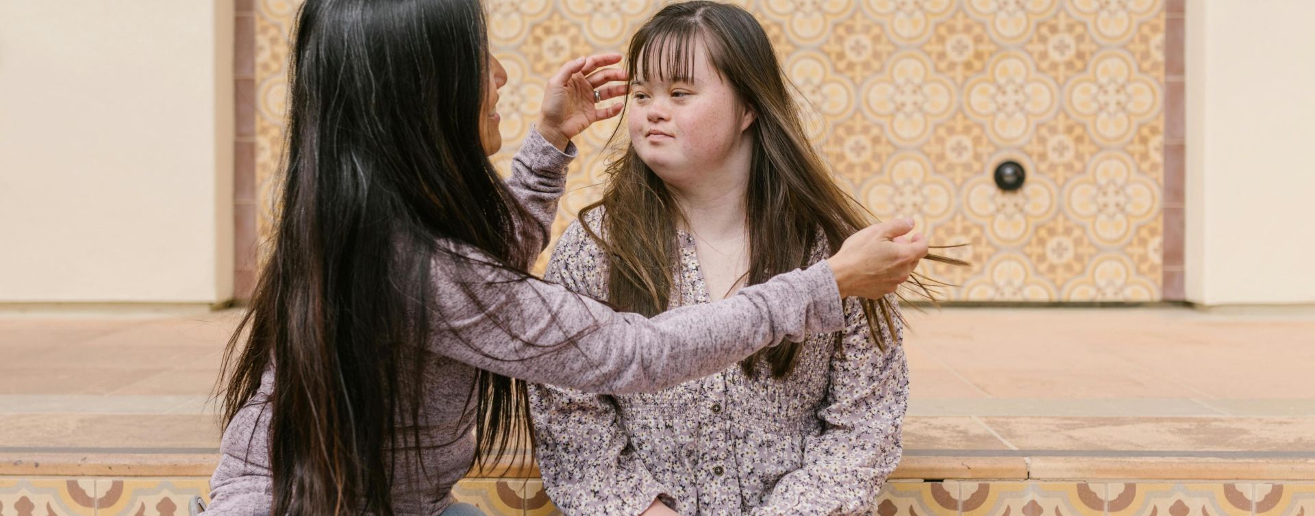 A mother lovingly adjusts her daughter's hair as they sit together on steps.