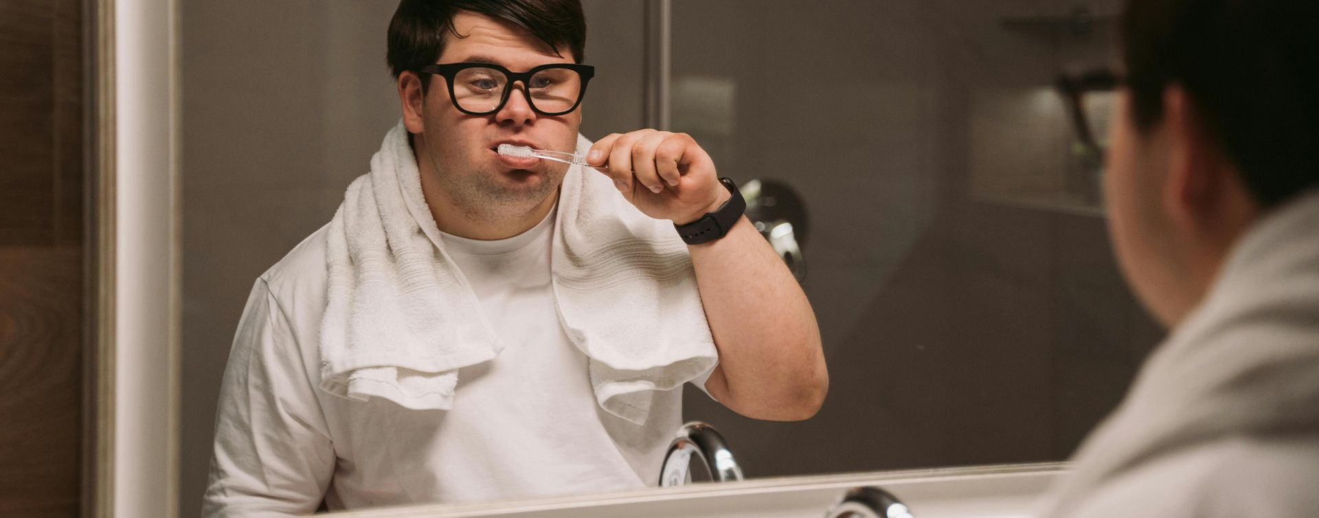 A man with Down syndrome brushes his teeth while looking in a bathroom mirror, wearing a towel and glasses.