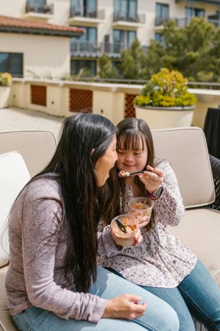 A mother and daughter with Down syndrome share a joyful moment eating pudding on a rooftop.